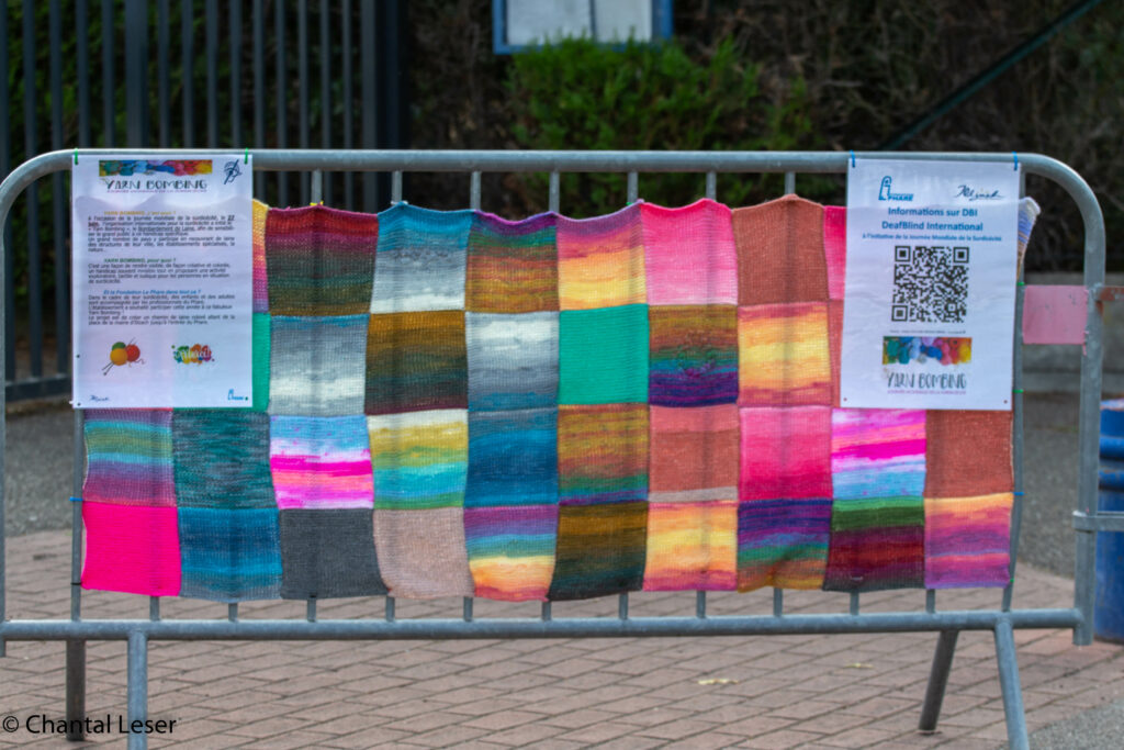 Plein de carré de laine assemblés et mis sur une barrière avec des affiches expliquant le Yarn bombing et la surdicécité