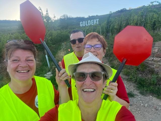 Carole, Chantal, Anne-Marie et Jean-Rémy devant les vignes, en tee-shirt rouge Vision'ère et gilet jaune. Anne-Marie tient les panneaux rouge de signalisation.