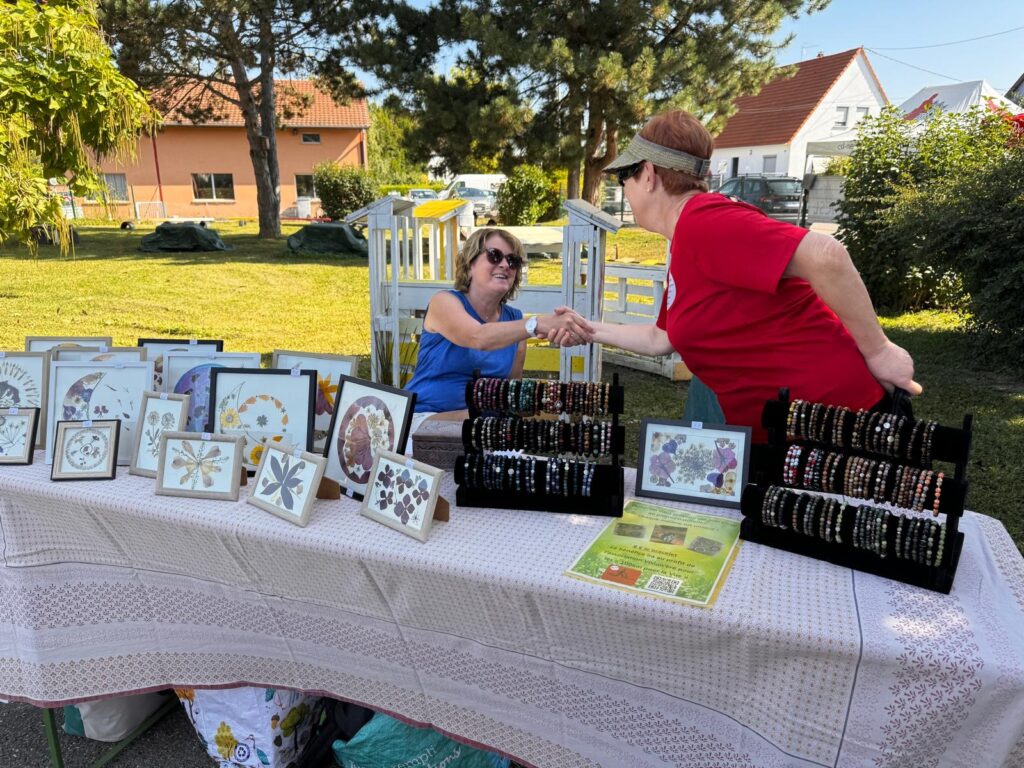 Catherine Gledel et Anne-Marie se saluent et en premier plan la table avec les bracelets et les tableaux que Catherine fabrique et vend au profit de Vision'ère