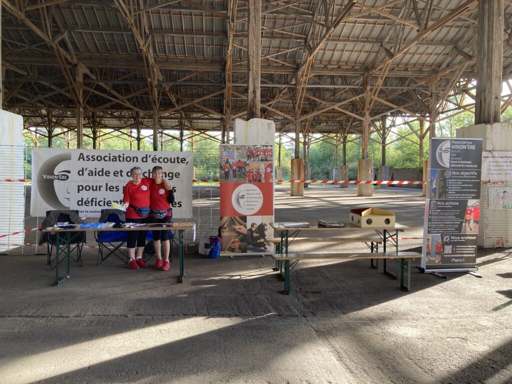 Vue du stand Vision'ère avec la banderole, les kakémonos, les jeux et le matériel pour la mise en situation. Rachel et Dominique prennent la pose